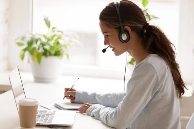A woman with brown hair, wearing a headset, is sitting at a desk in front of a laptop. Providing virtual assistance for lead generation, she's jotting notes in her notebook while sipping coffee. The backdrop showcases potted plants and a bright window.