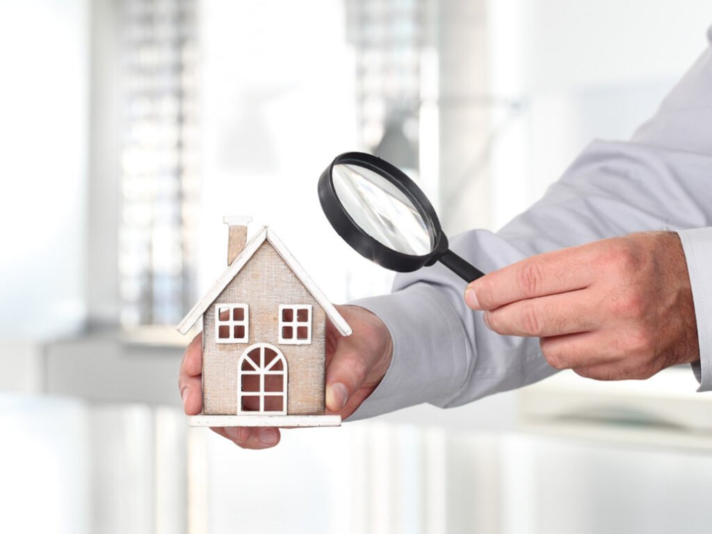 A person in a gray shirt examines a wooden model house with a magnifying glass in a bright, modern indoor setting.