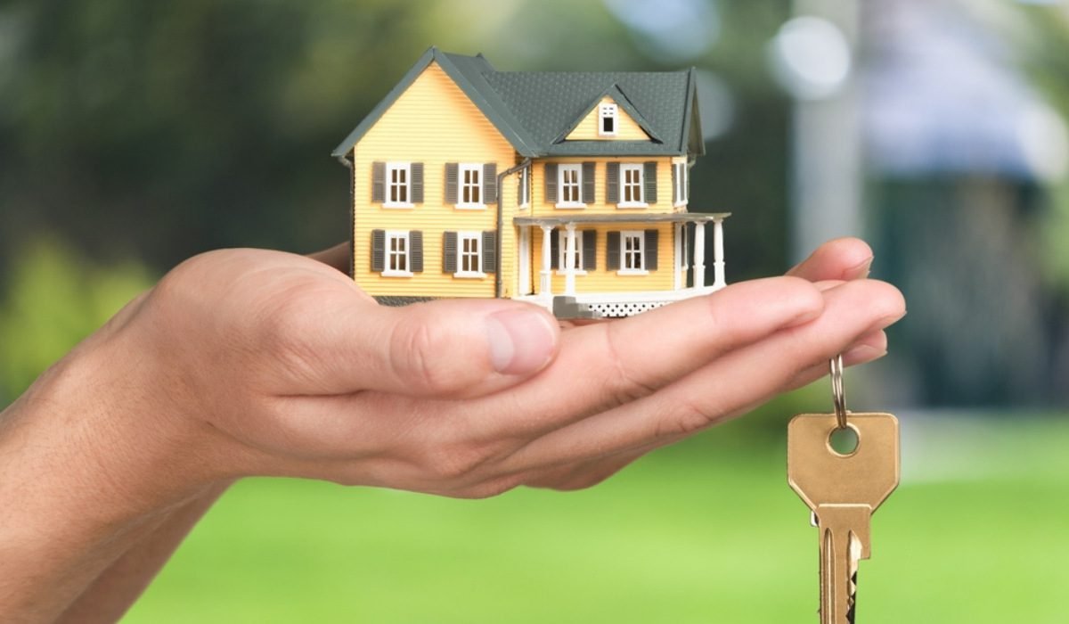 A hand holds a small model of a yellow house alongside a key, symbolizing home ownership and real estate.
