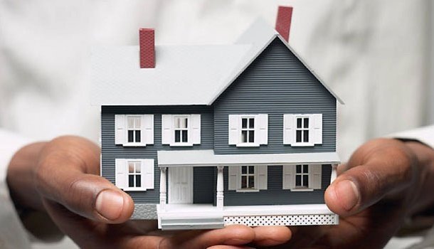 A person holds a small, detailed model of a gray house with white trim and red chimney, symbolizing homeownership or real estate.