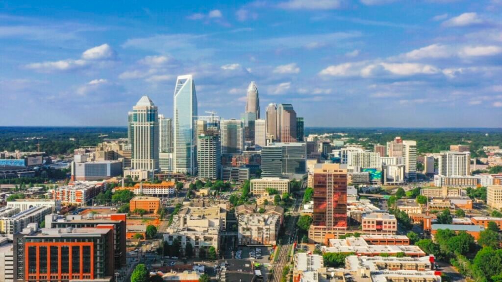 Aerial view of Charlotte's skyline featuring modern skyscrapers and a blue sky with fluffy clouds, surrounded by green space.