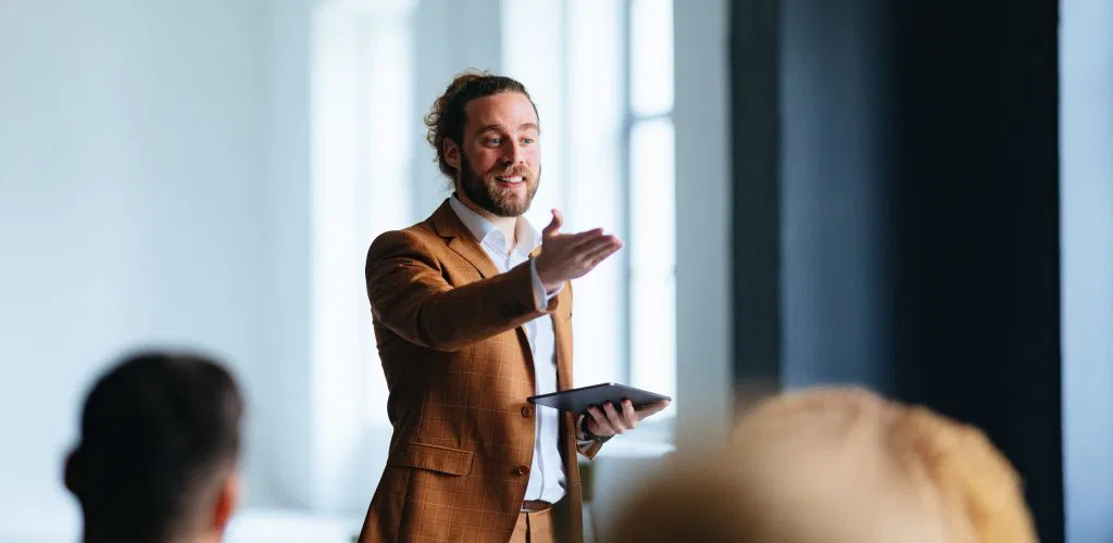A speaker in a brown suit gestures while holding a tablet during a presentation, with an audience visible in the foreground.