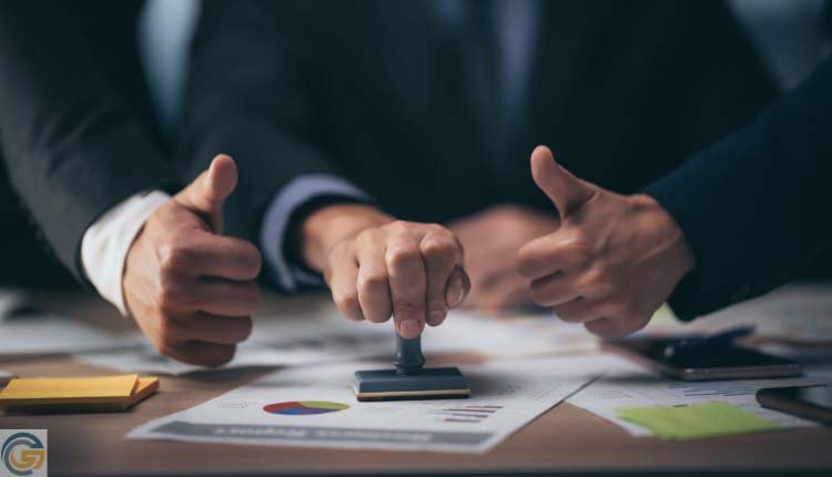 Three business professionals agree on a decision, giving thumbs up while one stamps a document on a table covered with reports and notes.