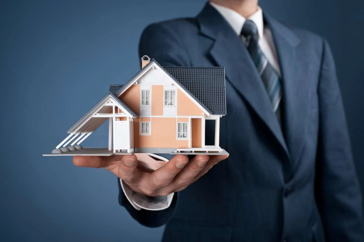 A businessman in a suit holds a detailed miniature house model in front of a blue background, symbolizing real estate or home buying.