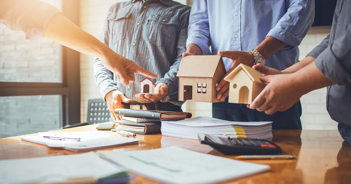 Four people holding miniature house models in a meeting, surrounded by papers and a calculator on a wooden table.