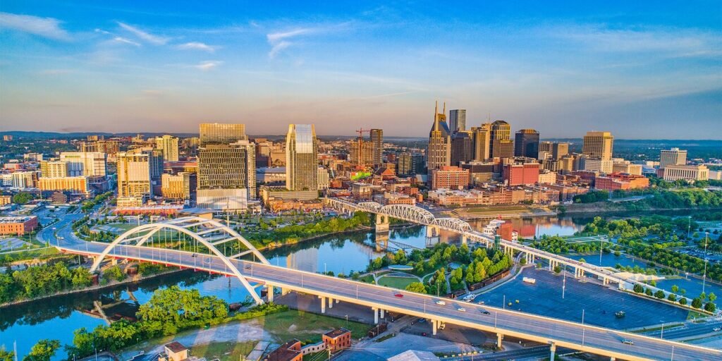 Aerial view of Nashville's skyline at sunset, showcasing rivers, bridges, and vibrant city buildings surrounded by greenery.