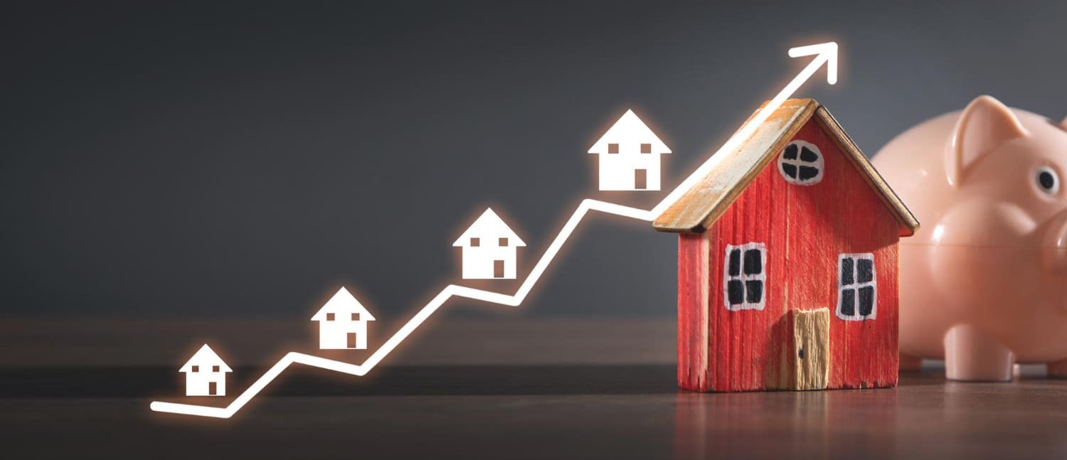 A wooden toy house next to a piggy bank, with homes and a rising graph line symbolizing a growing housing market.