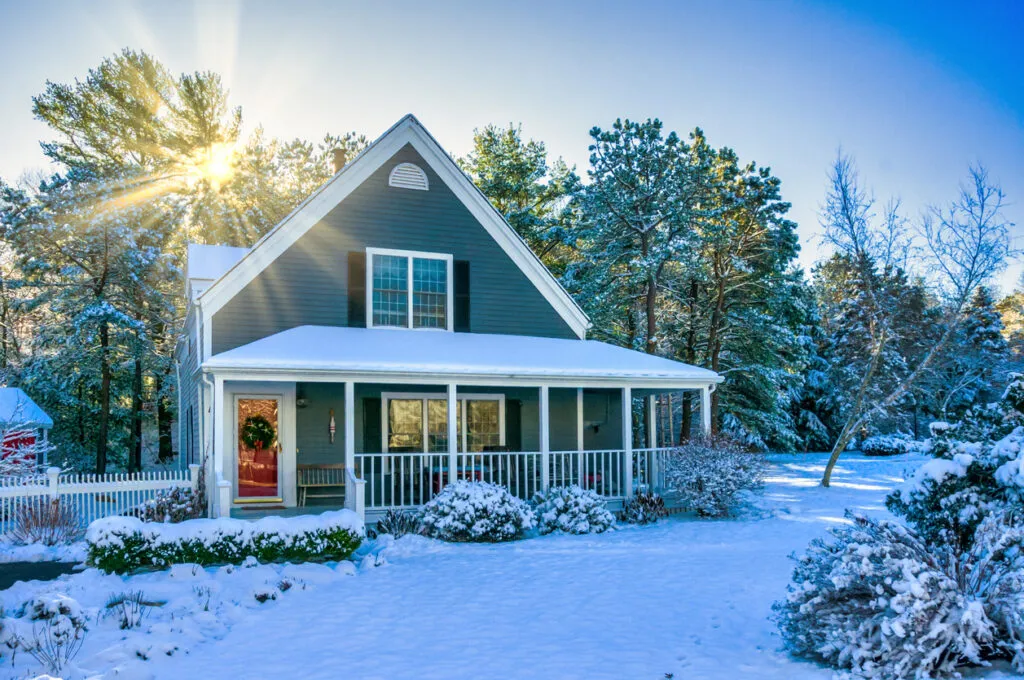 A charming gray house covered in snow, surrounded by trees, with sunlight shining through, creating a serene winter scene.