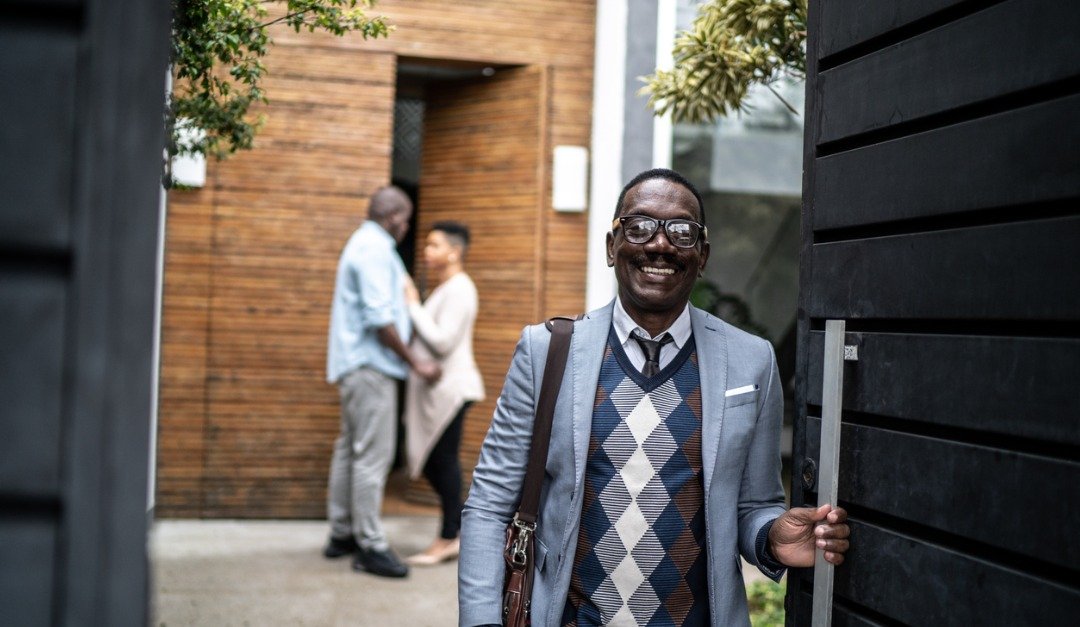 A professional real estate agent, dressed in a sweater and tie, poses in front of a house, showcasing his expertise in the market.