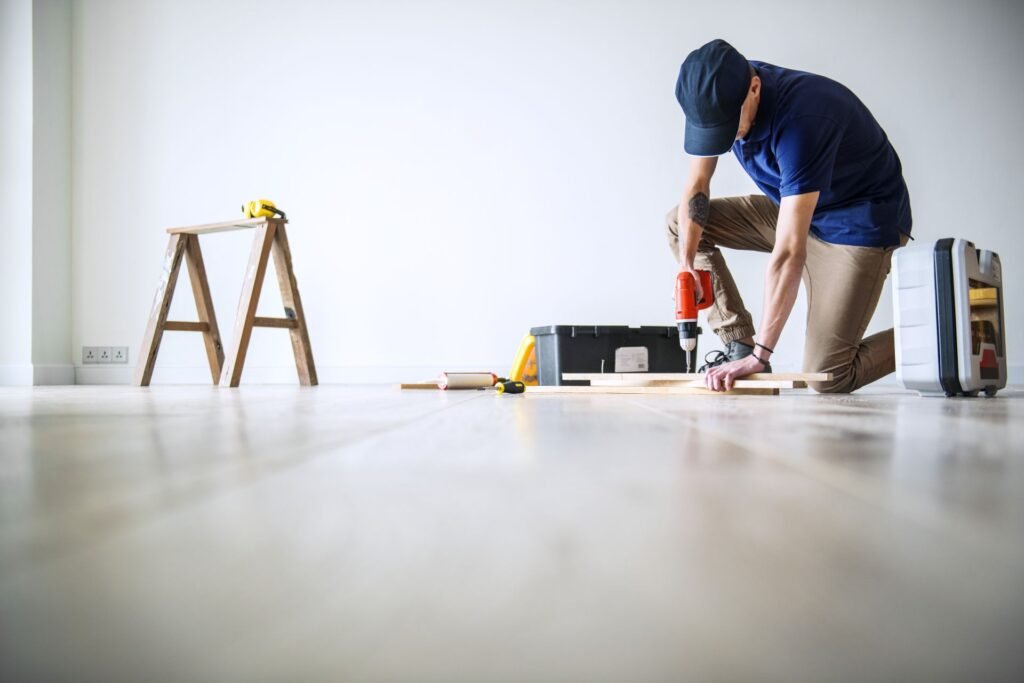 A person kneels on the floor using a red power drill on a wooden board, with tools and a ladder in a blank room.