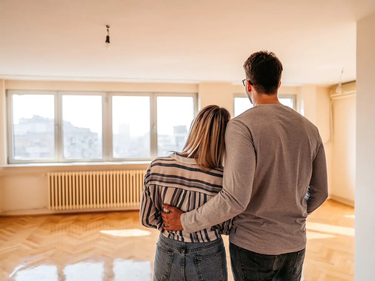 A couple stands together, gazing at an empty room with large windows, contemplating their future in the new space.