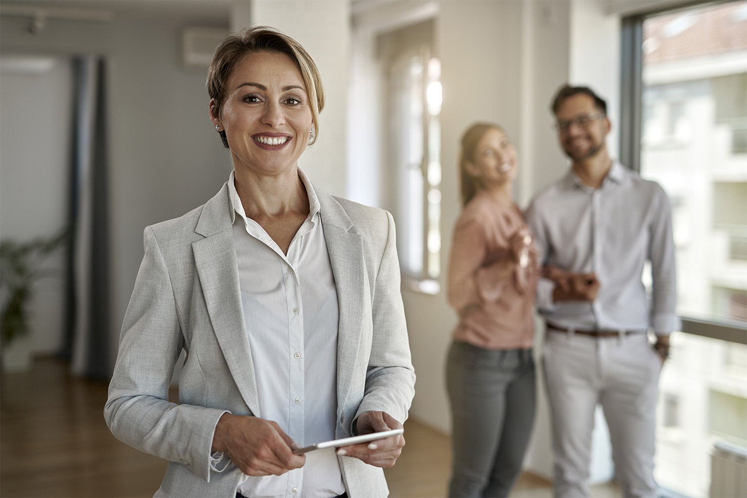 A professional woman in a light gray suit holds a tablet, while two colleagues converse in a bright office setting.