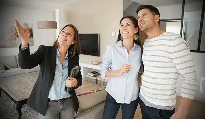 A woman in a black blazer gestures while showing a couple around a modern living room with neutral decor.