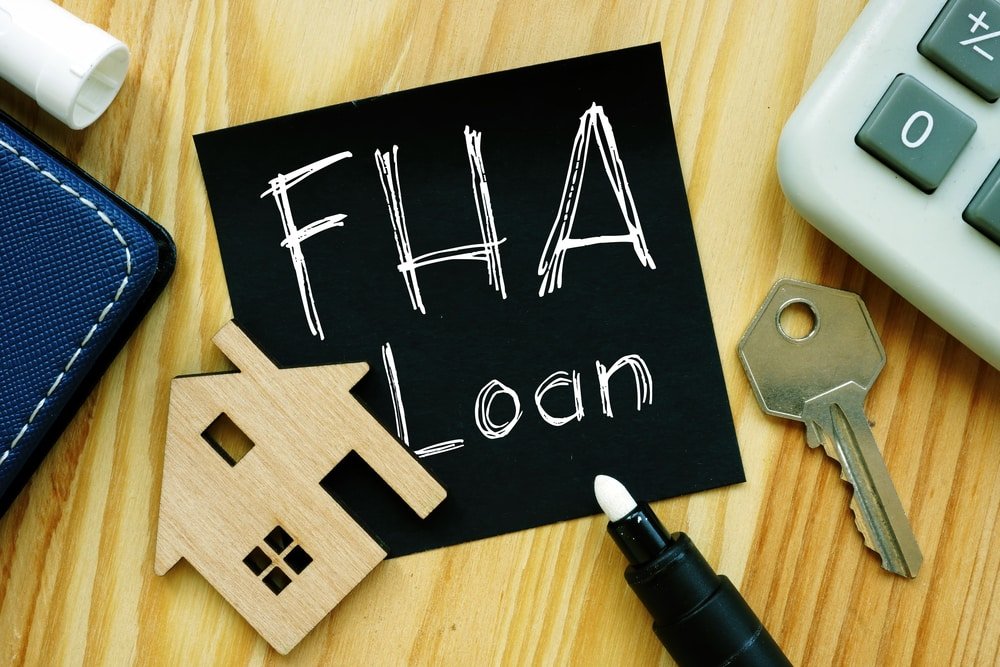Close-up of a sticky note labeled "FHA Loan," alongside a wooden house model, a key, a black marker, and a calculator on a wooden surface.