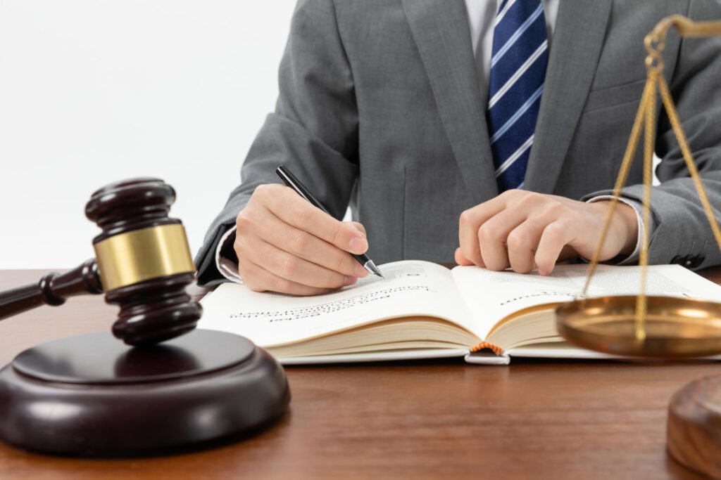 A suited lawyer writes notes in a legal book, with a gavel and scales of justice on the table beside him.