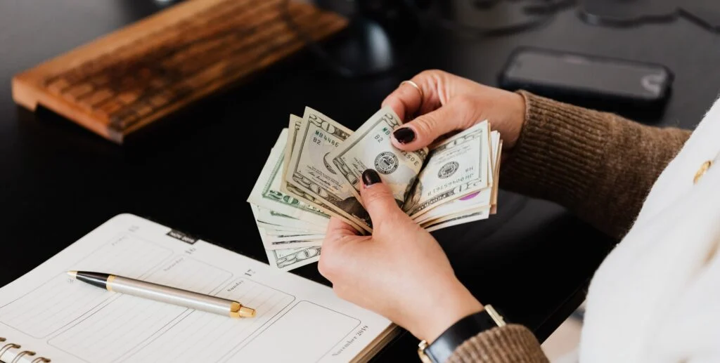 A person with dark nails counts a stack of cash while seated at a desk with a planner and a pen.