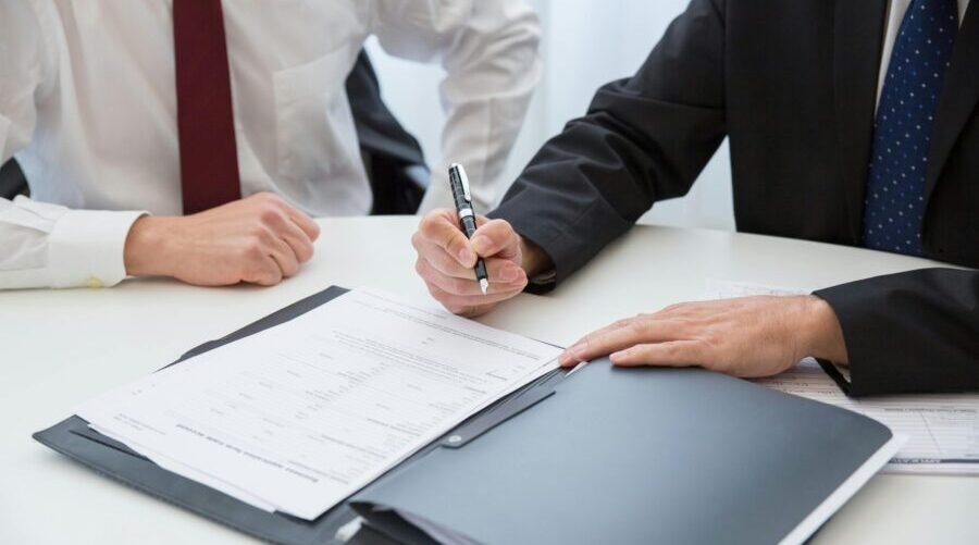 Two business professionals sit at a table, discussing documents. One man writes notes while the other observes attentively.