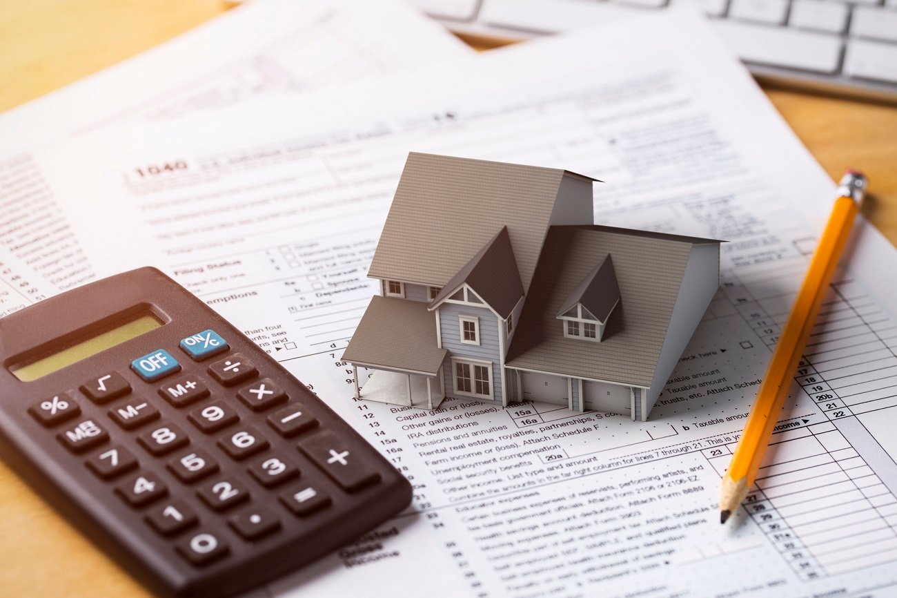 A calculator, a pencil, and tax documents surround a miniature house model, symbolizing real estate finances and tax preparation.