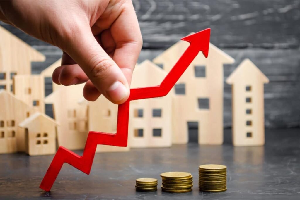 A hand holds a red upward arrow above stacks of coins, with wooden house models in the background, symbolizing economic growth.