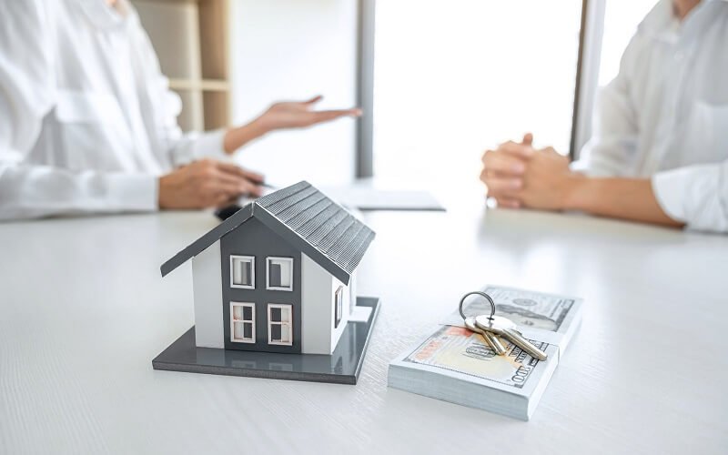 A miniature house on a table with cash and keys, while two people discuss real estate.