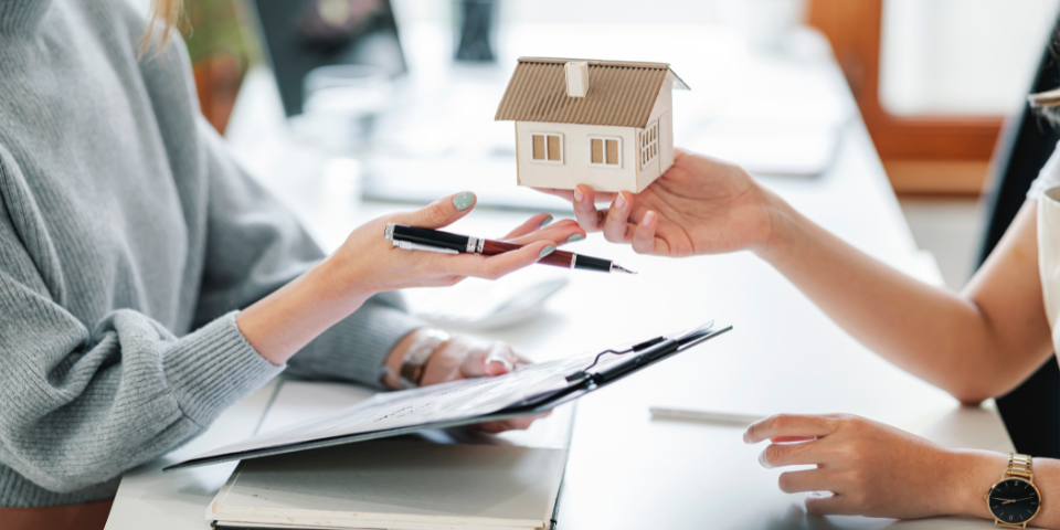 A woman hands a small model house to another woman at a desk, illustrating a real estate transaction.