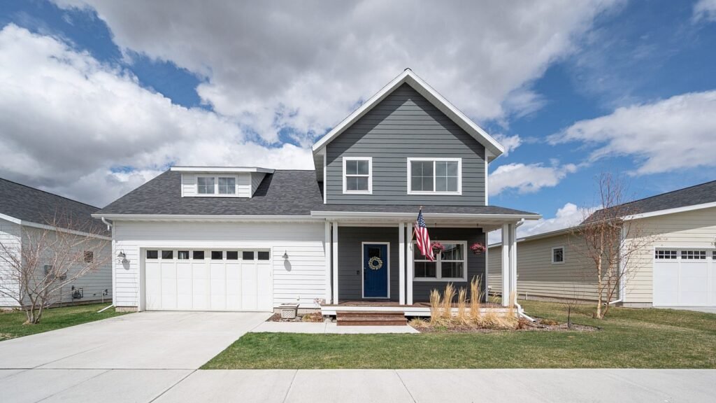 A modern two-story house with gray and white siding, an American flag, and a welcoming front porch, under a partly cloudy sky.