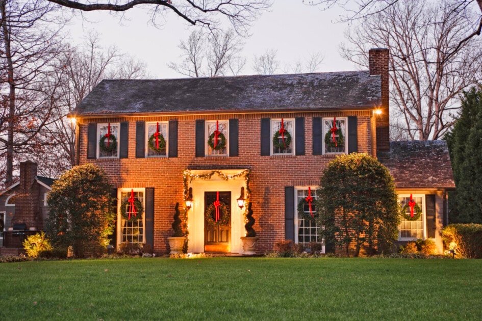 A charming two-story brick house adorned with holiday wreaths and lights, surrounded by a lush green lawn and barren trees at dusk.