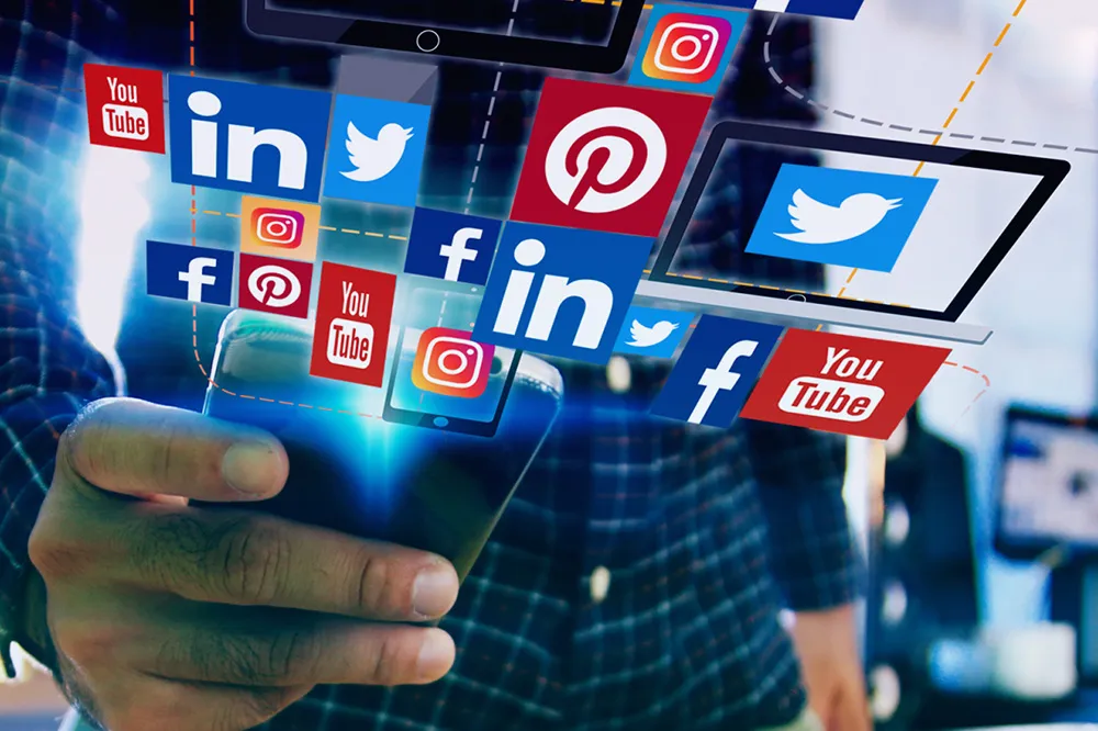 A person holding a smartphone with floating social media icons above, including YouTube, Twitter, Instagram, and LinkedIn.