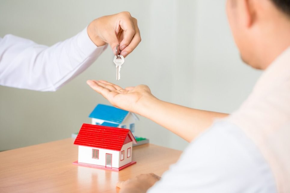 A person receives keys from another hand, with a small house model on a table, symbolizing a real estate transaction.