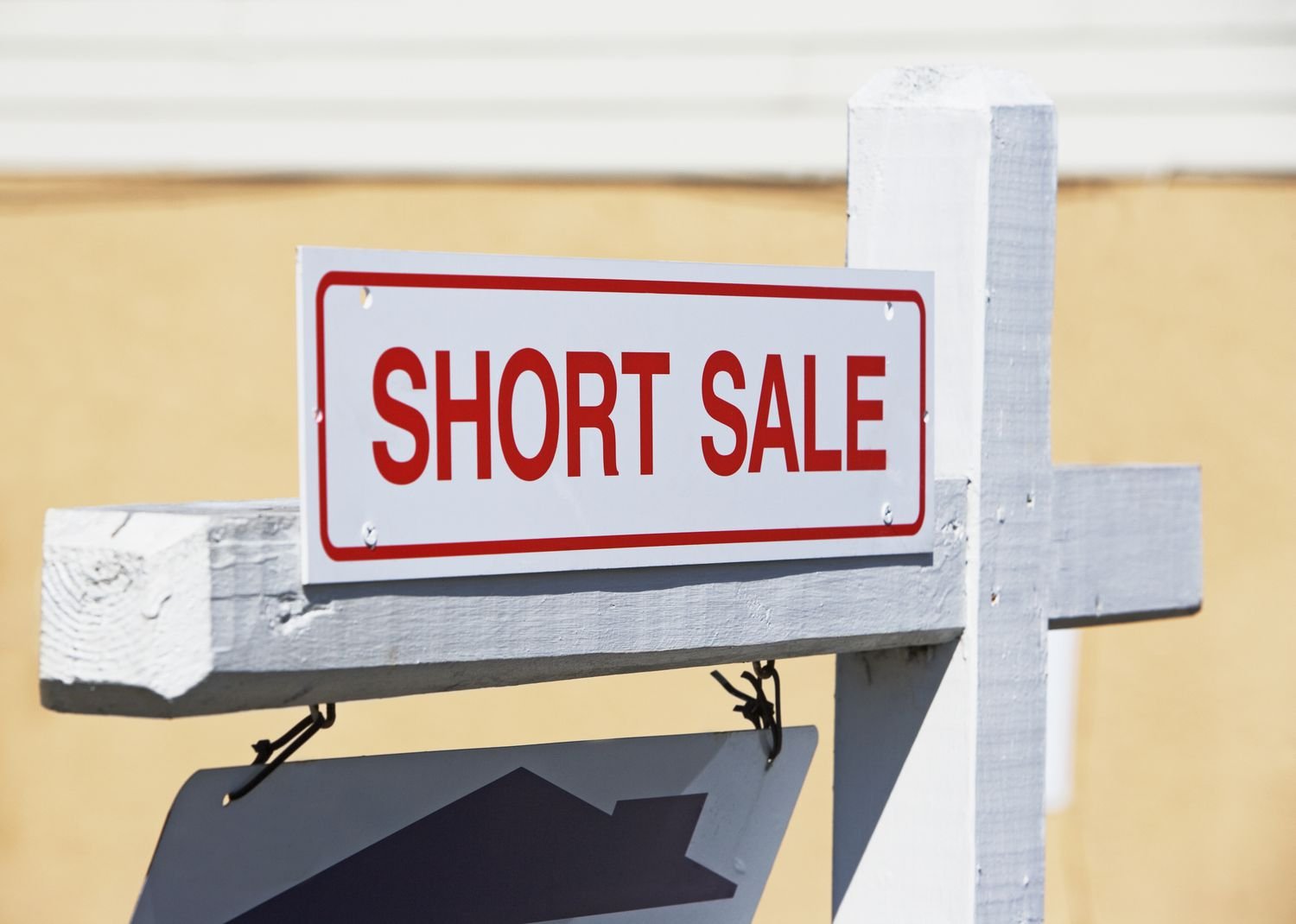 A real estate sign reading "SHORT SALE" in bold red letters, attached to a white wooden post against a beige background.