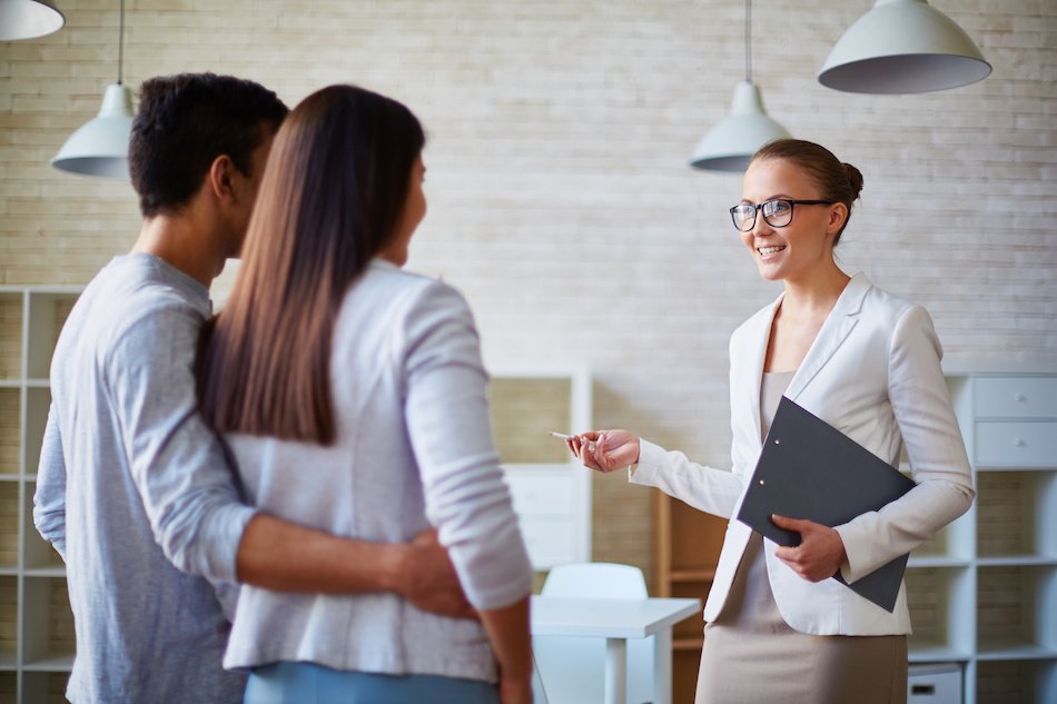A professional woman speaks to a couple, holding a folder and a pen, in a modern office setting with neutral decor.
