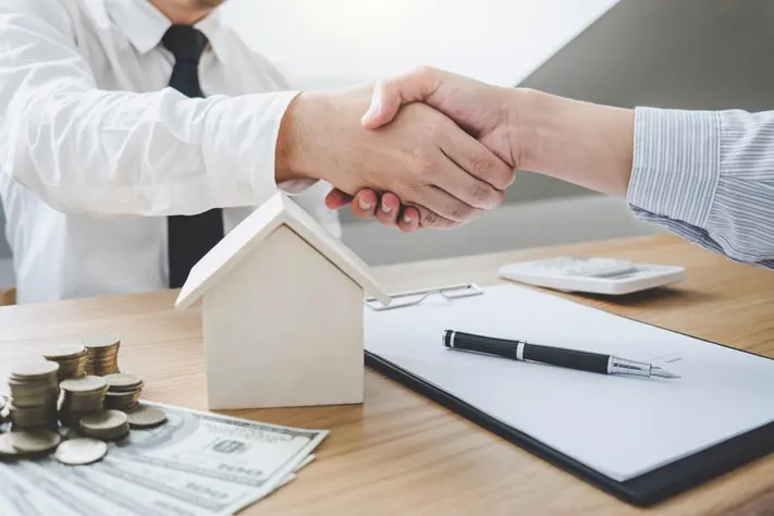 Two people shake hands over a desk with a small house model, money, and a contract, symbolizing a real estate agreement.