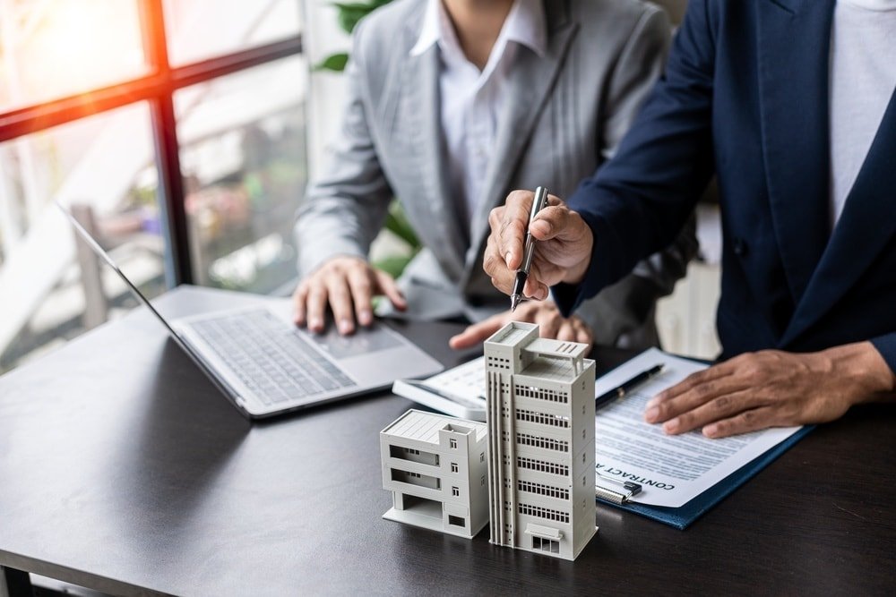Two business professionals signing a contract beside a laptop and a miniature building model on a desk. Bright windows in background.