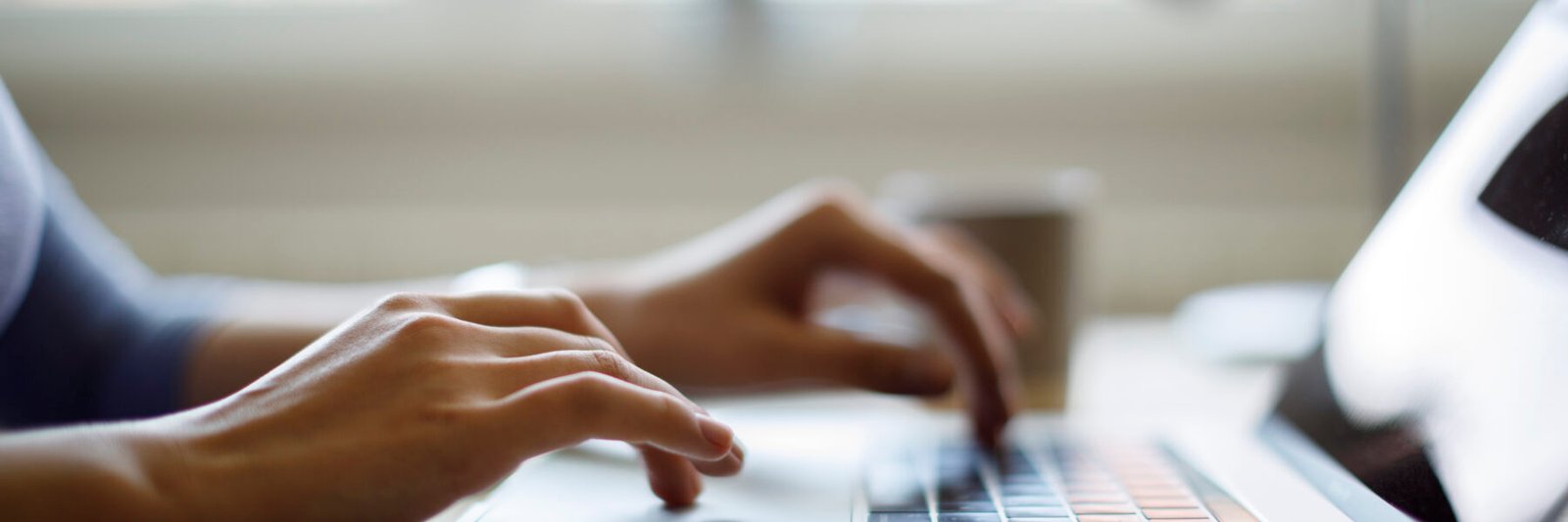 Close-up of a person’s hands typing on a laptop keyboard, perhaps brainstorming 25 summer social media ideas for real estate agents, with a blurred background featuring a coffee cup and sunlight streaming through the window.