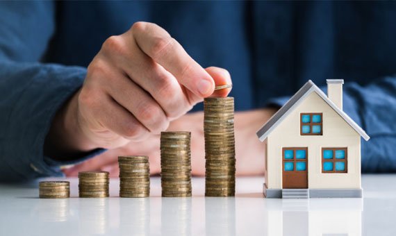 A person placing a coin on a stack of increasing coins next to a small model house, symbolizing financial growth and investment.