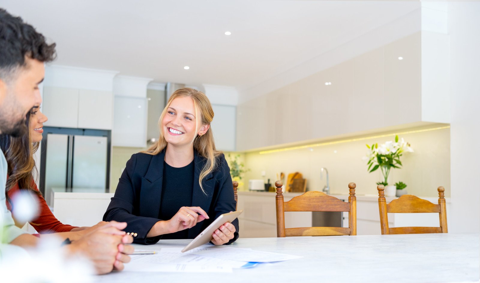 A smiling woman in business attire holds a tablet and discusses 9 Email Marketing Metrics Every Real Estate Agent Should Track with a couple seated at a table in a bright, modern kitchen, paperwork spread out before them.