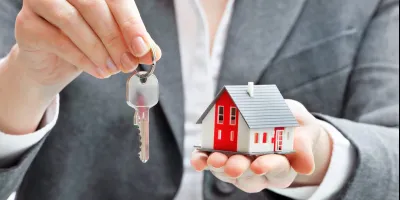 A person in a suit holds a house model and a key, symbolizing homeownership and real estate transactions.