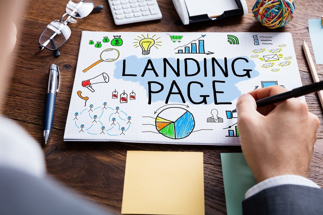 A person writes on a notepad with LANDING PAGE in bold letters, surrounded by colorful business icons and graphs, illustrating How to Optimize Your Landing Page for More Leads on a wooden desk with office supplies like a calculator and sticky notes.