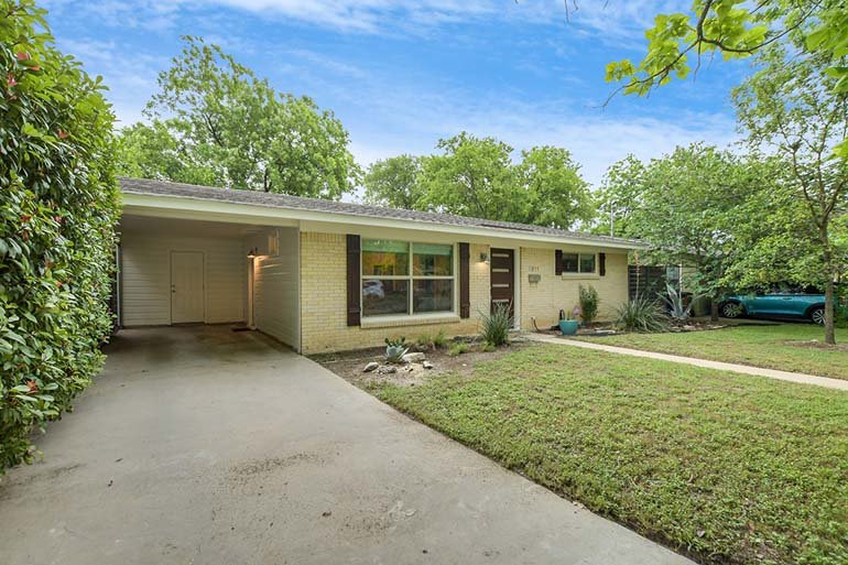 Single-story home with a manicured lawn, driveway, and surrounded by greenery under a blue sky. A turquoise car is parked nearby.