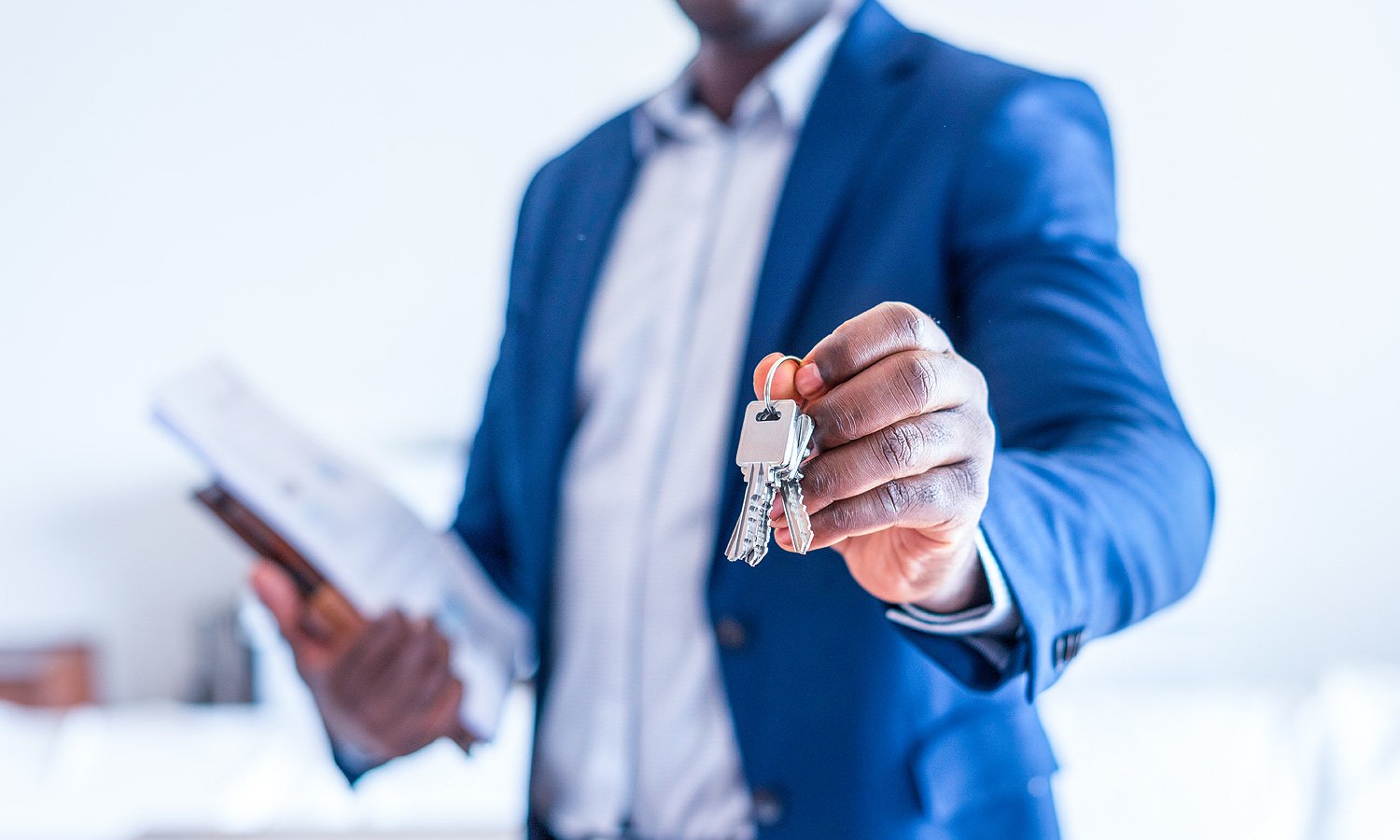 A man in a navy suit holds out a set of keys in one hand and a document in the other, set against a bright background.