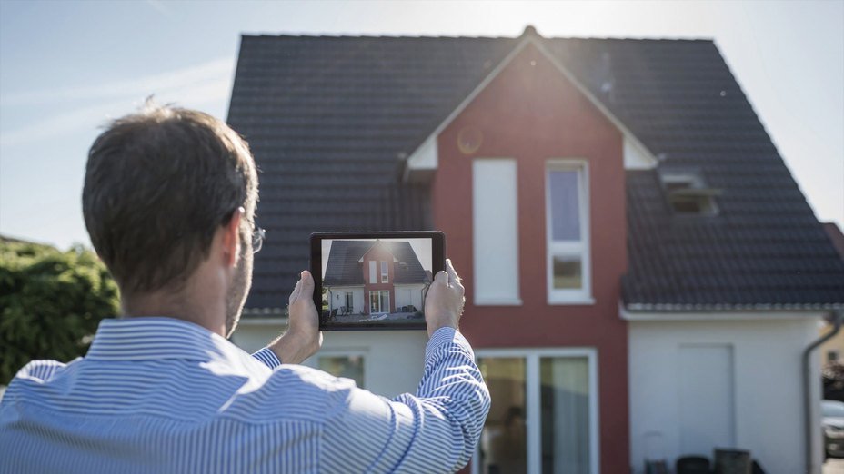 A man in a striped shirt captures a photo of a red and gray house using a tablet under bright sunlight.