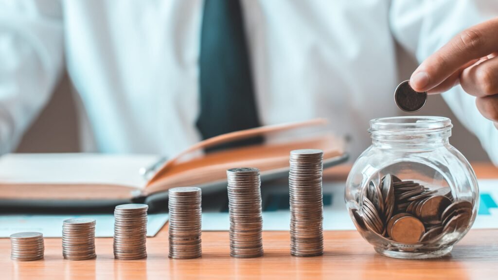 A person in a white shirt and black tie adds a coin to a jar amidst stacks of coins and an open notebook on a desk.