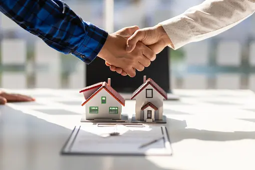 Two individuals shake hands over a table with miniature houses and paperwork, symbolizing a real estate agreement.