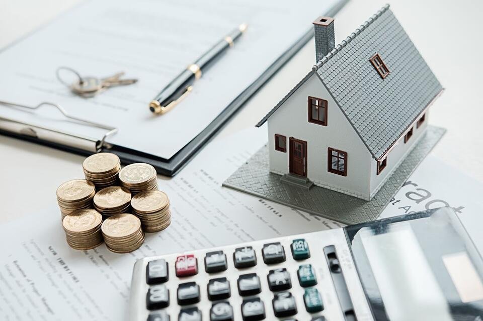 A small model house surrounded by coins, a calculator, and paperwork, symbolizing real estate and financial planning.