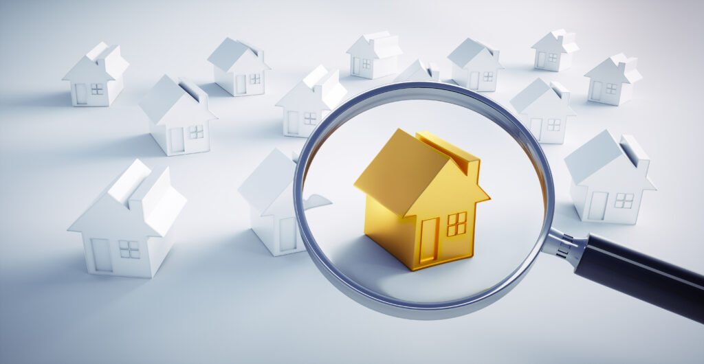 A magnifying glass focuses on a shiny gold house among a cluster of white miniature houses, symbolizing real estate selection.