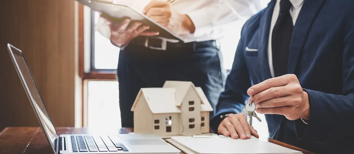 A businessman hands over keys while discussing real estate, with a laptop and a model house on a desk in a well-lit office.