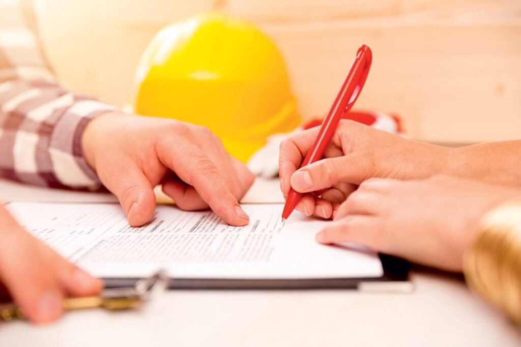 Two hands signing documents with a red pen, a yellow hard hat in the background, and a focus on collaboration and agreement.