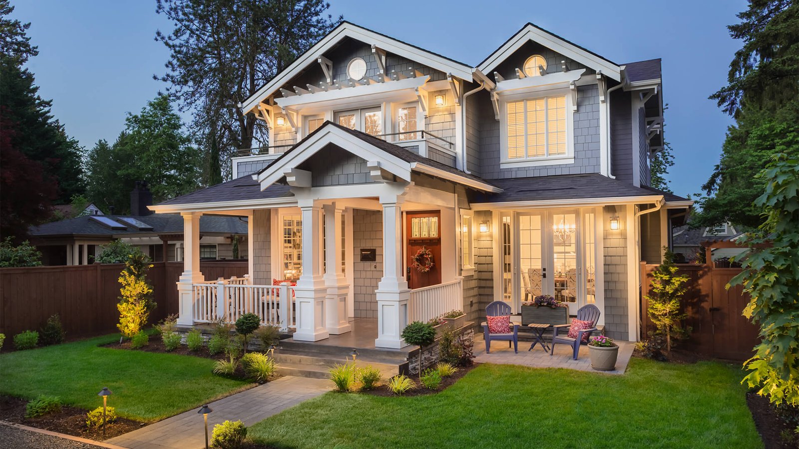 Two-story house with gray siding and white trim, front porch, outdoor seating area, landscaped yard, and lights on at dusk.