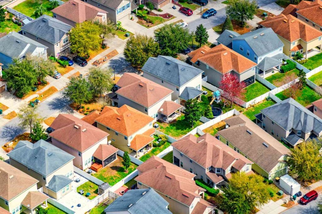 Aerial view of a suburban neighborhood featuring houses with orange roofs, green lawns, and winding streets lined with trees and cars.