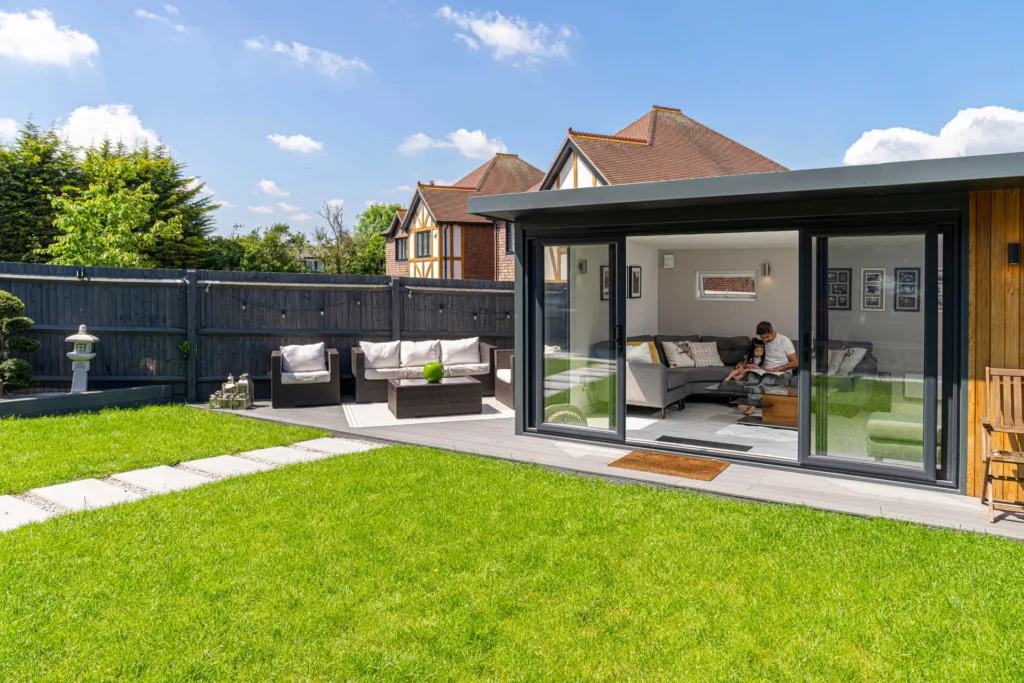 A bright backyard with lush green grass, modern outdoor seating, and a cozy interior visible through glass doors.